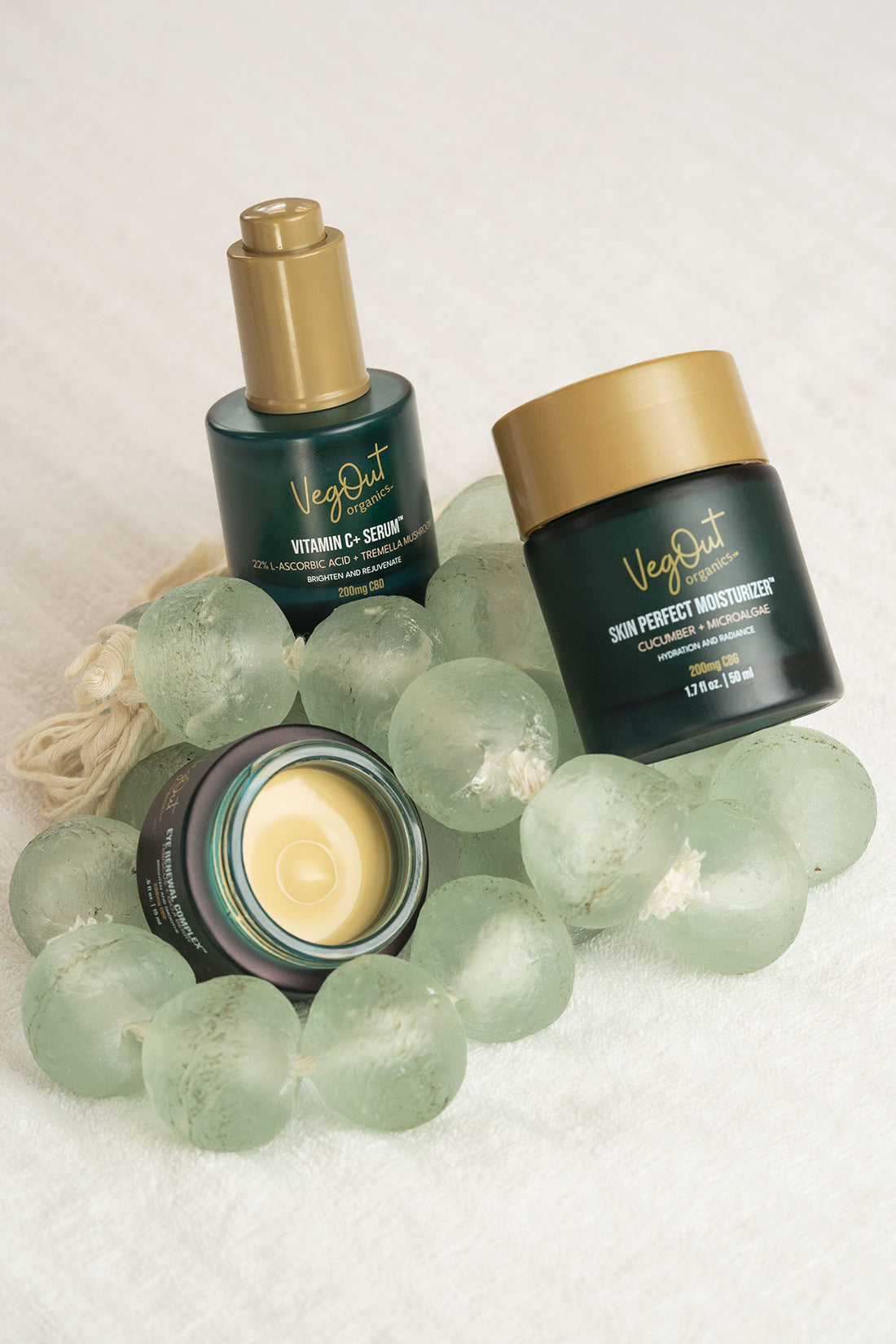 Flat lay of VegOut Organics skincare trio: Vitamin C+ Serum, Skin Perfect Moisturizer, and Eye Renewal Complex, surrounded by translucent glass beads on a soft towel background – natural skincare for hydration, radiance, and rejuvenation.
