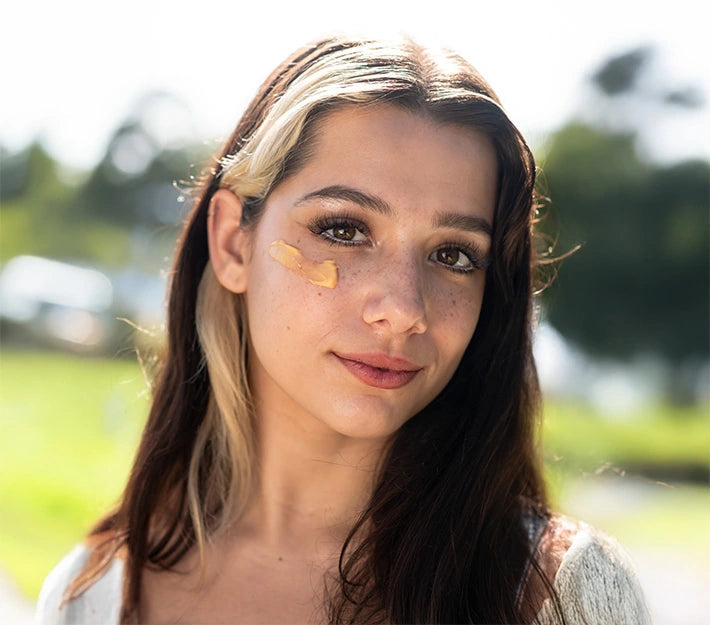 Young woman outdoors wearing a pumpkin-colored facial mask under her eye, promoting natural skincare and radiant complexion.