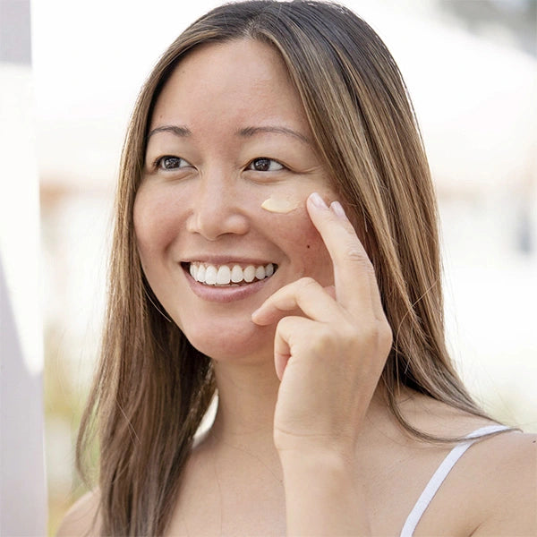 Smiling woman applying vegan eye cream to reduce fine lines and hydrate under-eye area naturally.